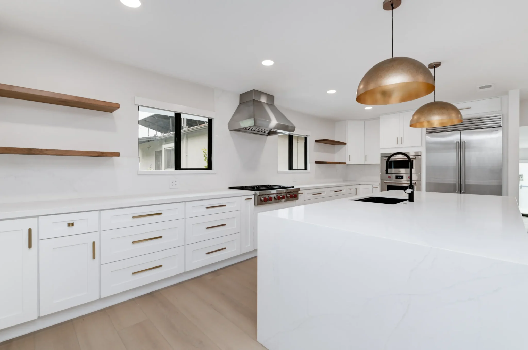 A modern white kitchen featuring white shaker-style cabinets with gold hardware, white quartz countertops, and light wood floating shelves on the left wall. The space includes a professional-grade stainless steel range with red knobs under a metal hood, stainless steel refrigerator, and a large white island with black undermount sink. Two copper-toned dome pendant lights hang over the island, complementing the warm wood flooring.