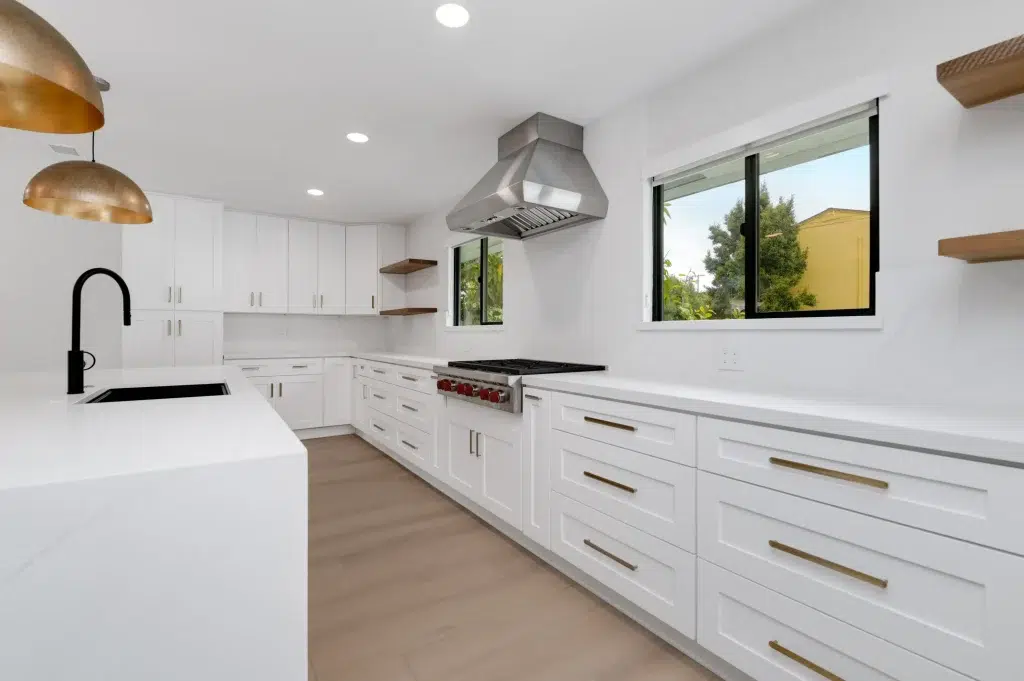 A modern L-shaped kitchen featuring crisp white shaker-style cabinets with gold hardware and white quartz countertops. The space includes a professional stainless steel range with matching hood, black matte faucet, floating wood shelves, and gold pendant lights hanging over a white island. Two windows with black frames provide natural light and views of the surrounding greenery, while light wood flooring completes the clean, contemporary design.