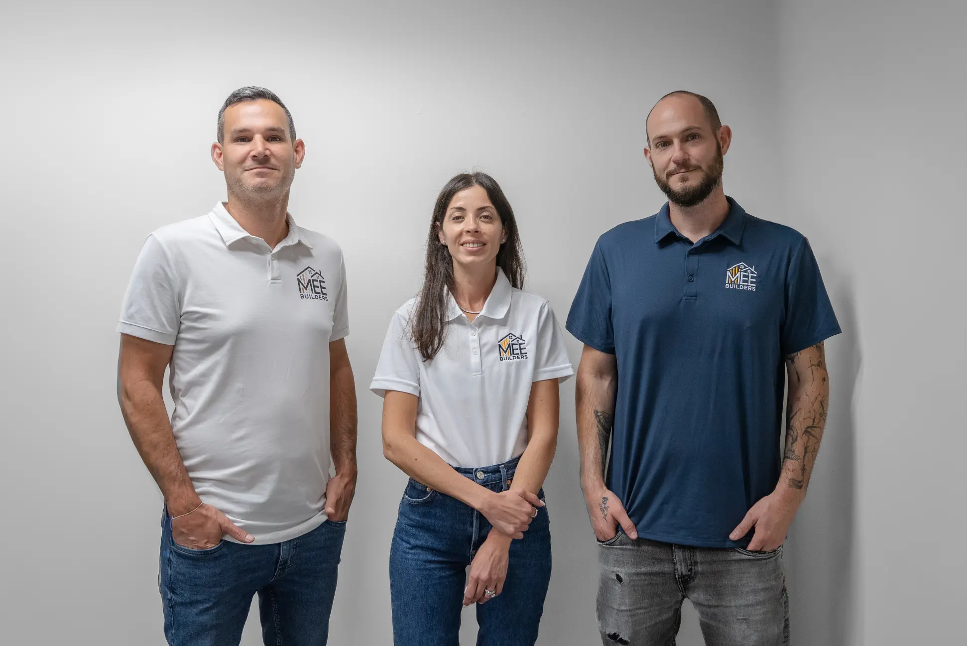 Three professionals from MEE Builders standing side by side against a white wall. The two men wear polo shirts in white and navy blue respectively, while the woman in the center wears a white polo shirt. All three are dressed in jeans and their shirts display the MEE Builders company logo.