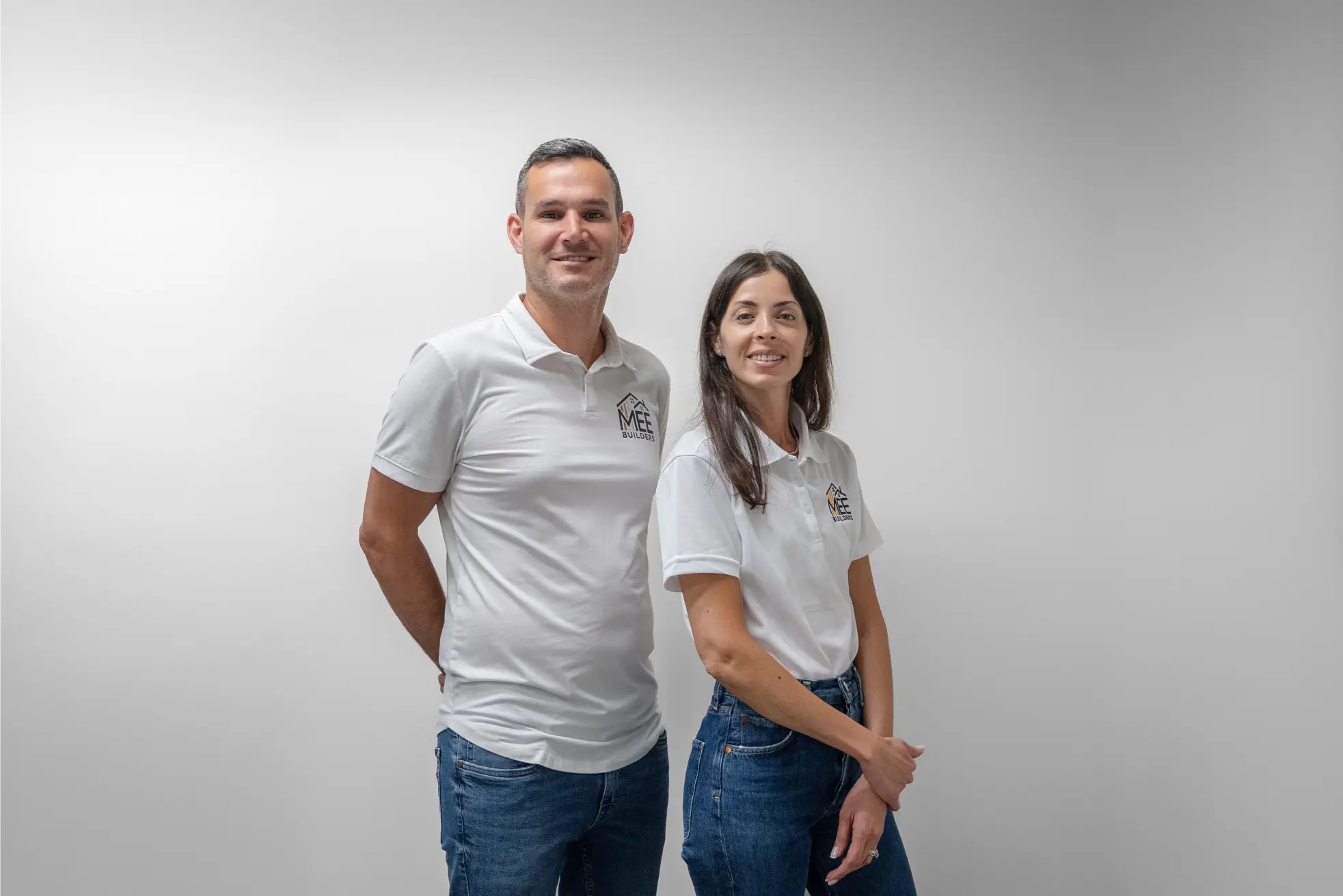 Two people standing side by side wearing matching white polo shirts with an "MEE BUILDERS" logo and blue jeans against a plain white background. The man on the left and woman on the right are both smiling at the camera in what appears to be a professional business portrait for their building or construction company.