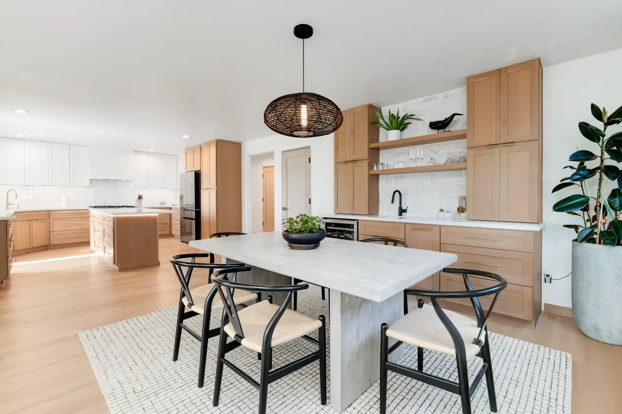A modern open-concept kitchen featuring light natural wood cabinets paired with white upper cabinets and marble countertops. The space includes a large white kitchen island with three black wishbone-style chairs, topped by a black woven pendant light. The kitchen showcases clean lines, open shelving, black fixtures, and is accented with potted plants including a tall fiddle leaf fig tree.