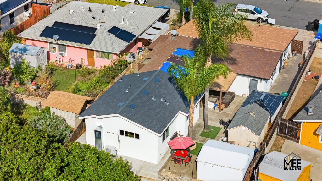 Aerial view of a residential neighborhood showing several single-family homes with different colored roofs including dark gray, brown, and light gray shingles. The properties feature solar panels on some roofs, palm trees, backyard patios with outdoor furniture including a red umbrella, and small yards with fencing. The image appears to be a professional real estate or construction photography shot taken by MEE Builders, as indicated by the watermark.