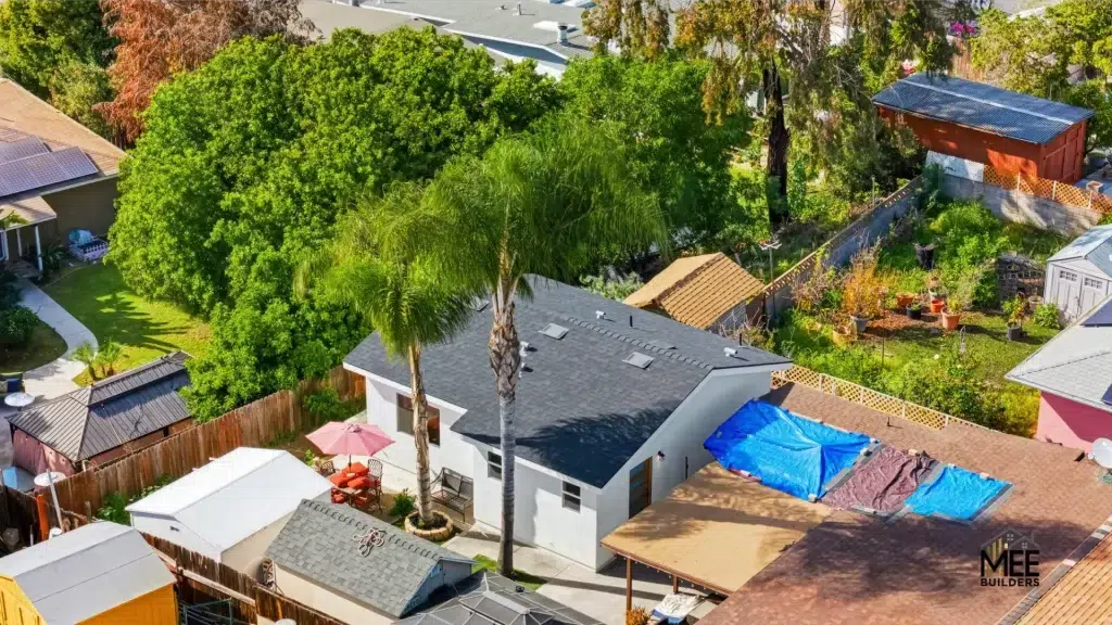 Aerial view of a residential neighborhood showing a house with a dark gray roof and white exterior, surrounded by lush green trees including tall palm trees. A blue tarp is visible on an adjacent brown-tiled roof, likely covering repair work. The property features wooden fencing and is surrounded by other homes with solar panels, garages, and well-maintained landscaping in what appears to be a suburban California setting.