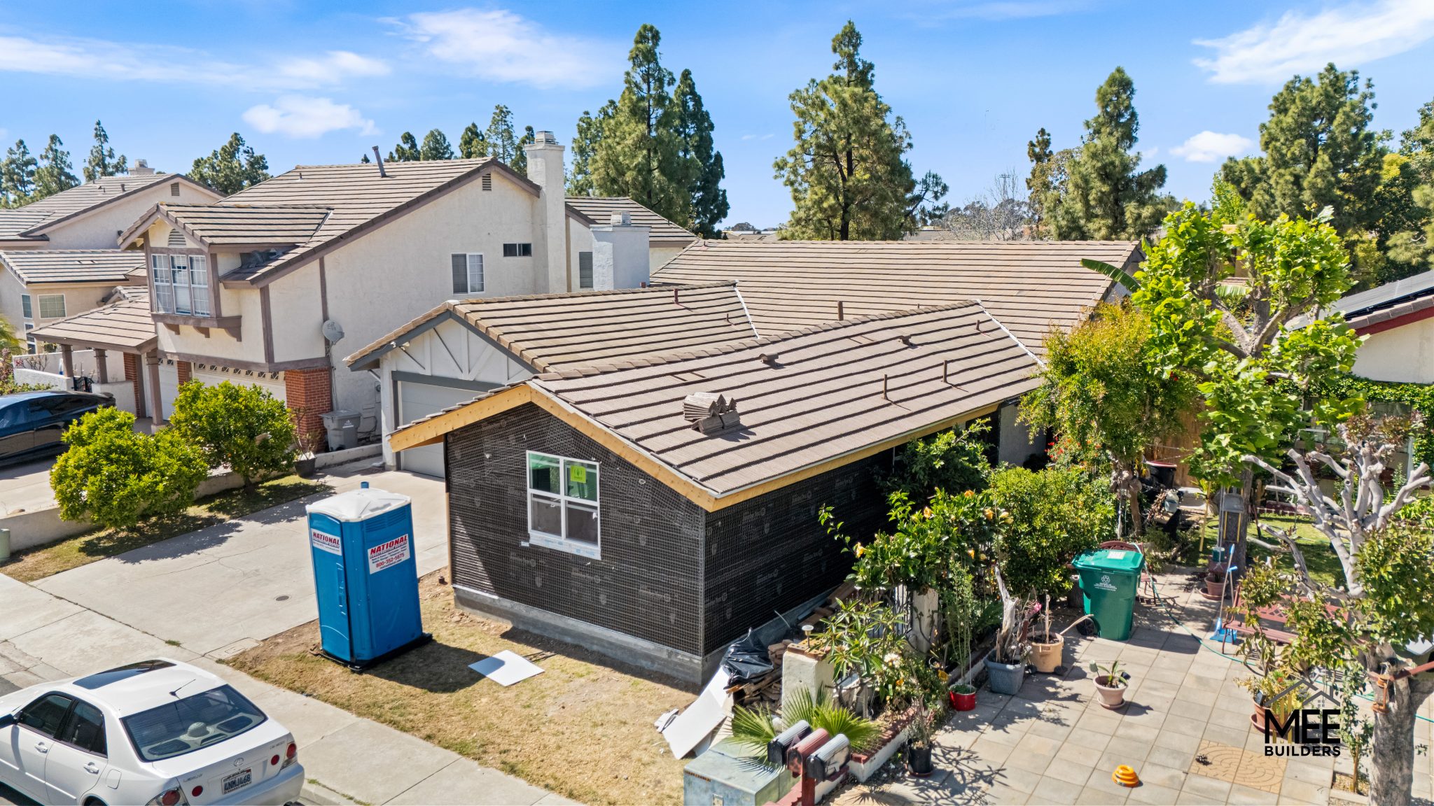 Aerial view of a single-story residential home with dark brick exterior and brown tile roofing under renovation, with a blue portable toilet in the driveway. The house is situated in a suburban neighborhood with mature trees and similar homes visible in the background. A white car is parked on the street and various plants and containers are visible in the front yard.
