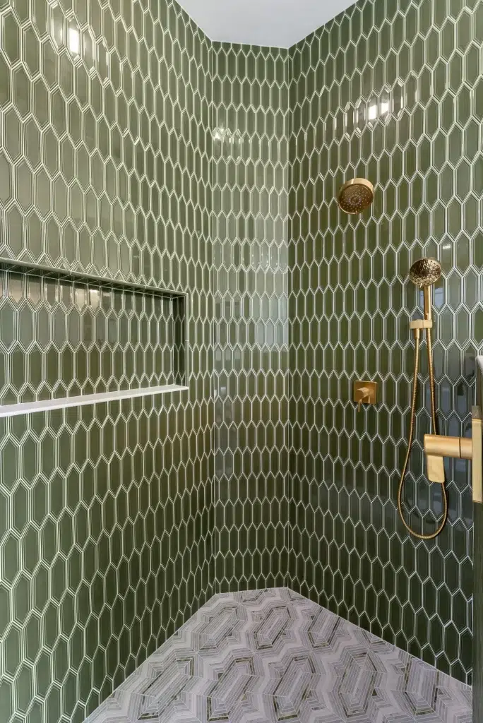 A modern shower interior featuring elongated hexagonal tiles in rich sage green covering the walls, complemented by brass-finished fixtures including a rainfall showerhead and handheld shower attachment. The floor displays an intricate geometric pattern in neutral gray tones with a three-dimensional cube effect. A built-in recessed shelf niche with white interior provides storage space within the green tiled wall.