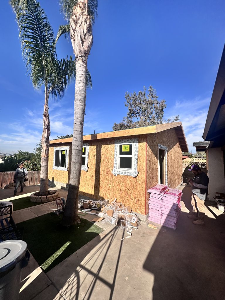 A two-story residential structure under construction with exposed oriented strand board (OSB) sheathing and window openings. The building features a peaked roof and is photographed from ground level with palm trees and a clear blue sky in the background. Construction materials including pink insulation boards are visible on the ground near the building.