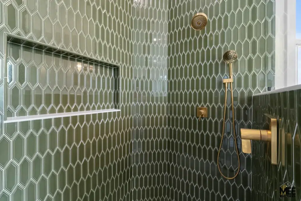 A modern shower wall featuring distinctive elongated hexagonal tiles in sage green with white geometric outlines creating an Art Deco-inspired pattern. Brushed gold or brass bathroom fixtures including a rainfall showerhead, handheld shower wand, and control valve are mounted on the tiled wall. A horizontal wall niche for storage is integrated into the tile design on the left side.