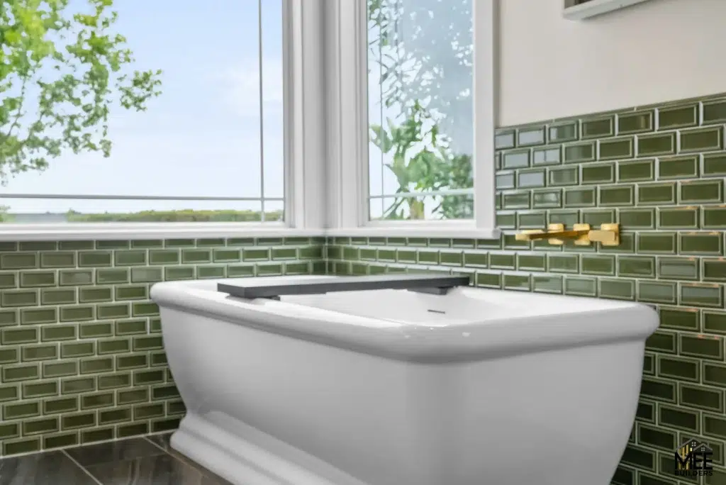 A modern bathroom featuring a white freestanding bathtub positioned against an olive green subway tile wall. Large windows above the tub reveal leafy trees and a peaceful outdoor view. The contemporary design includes gold fixtures and a clean, minimalist aesthetic with natural lighting.