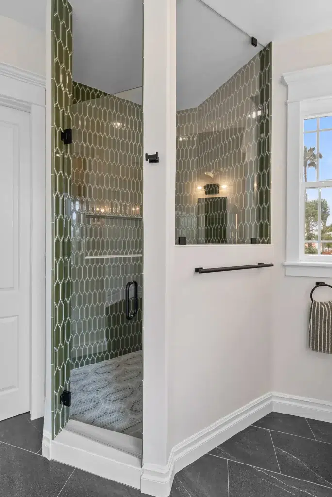 A modern bathroom shower featuring olive green elongated hexagonal tiles covering the walls, with a clear glass door and black hardware. The shower includes a built-in wall niche and mounted shelf, while the floor displays a gray herringbone tile pattern. White walls and black marble-look floor tiles complete the contemporary design outside the shower enclosure.