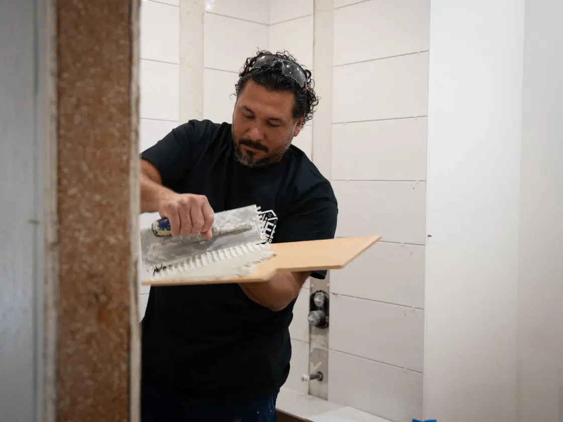 Tile installer back-buttering a tile with a notched trowel and thin-set adhesive in a San Diego spa bathroom renovation.