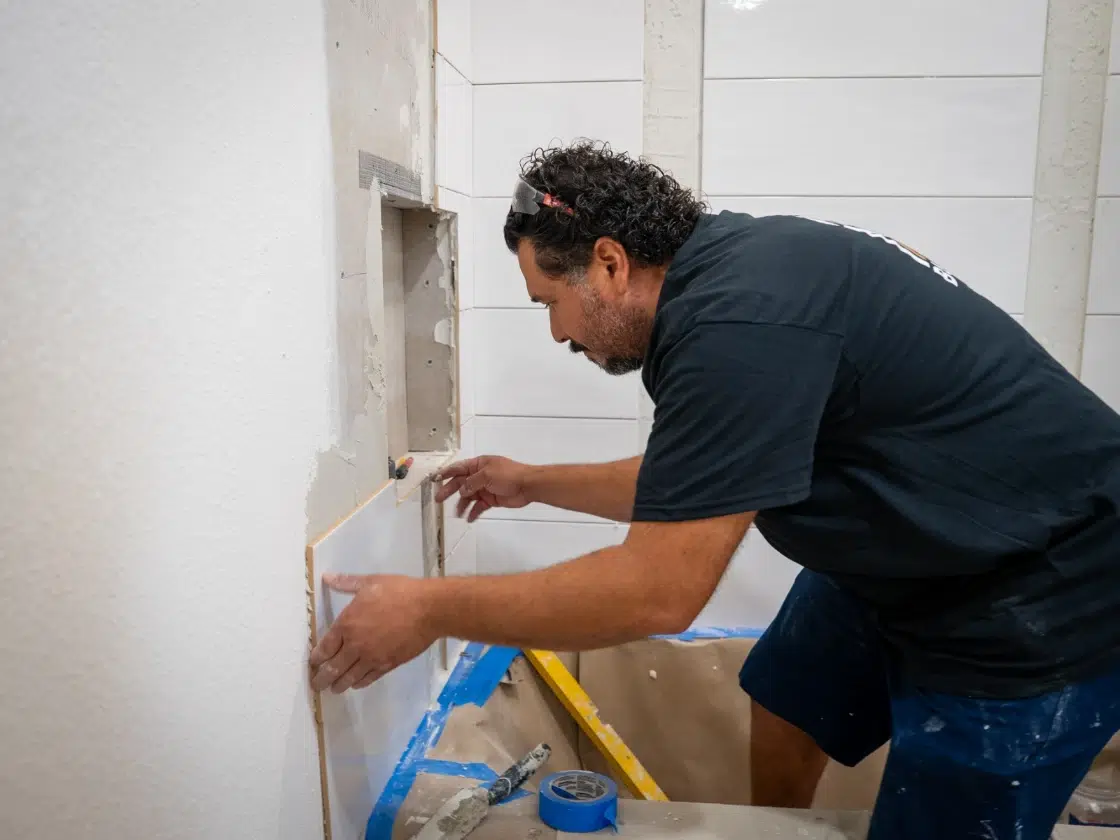 Installer setting custom white wall tile over sheet-membrane waterproofing with exposed rough-in plumbing, San Diego bathroom remodel.