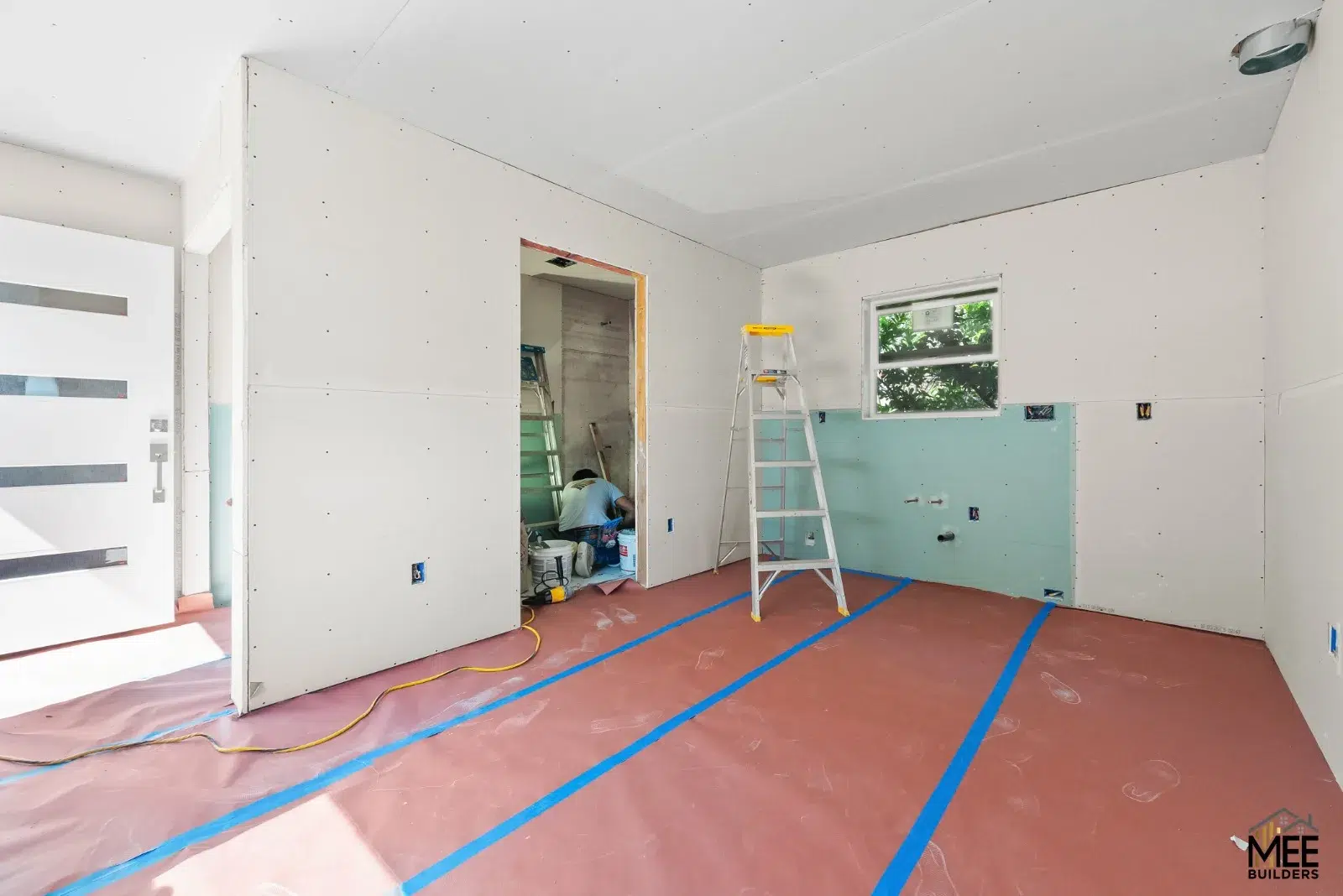 Unfinished drywall room with a stepladder, green wall panels, and red floor protection secured with blue tape.