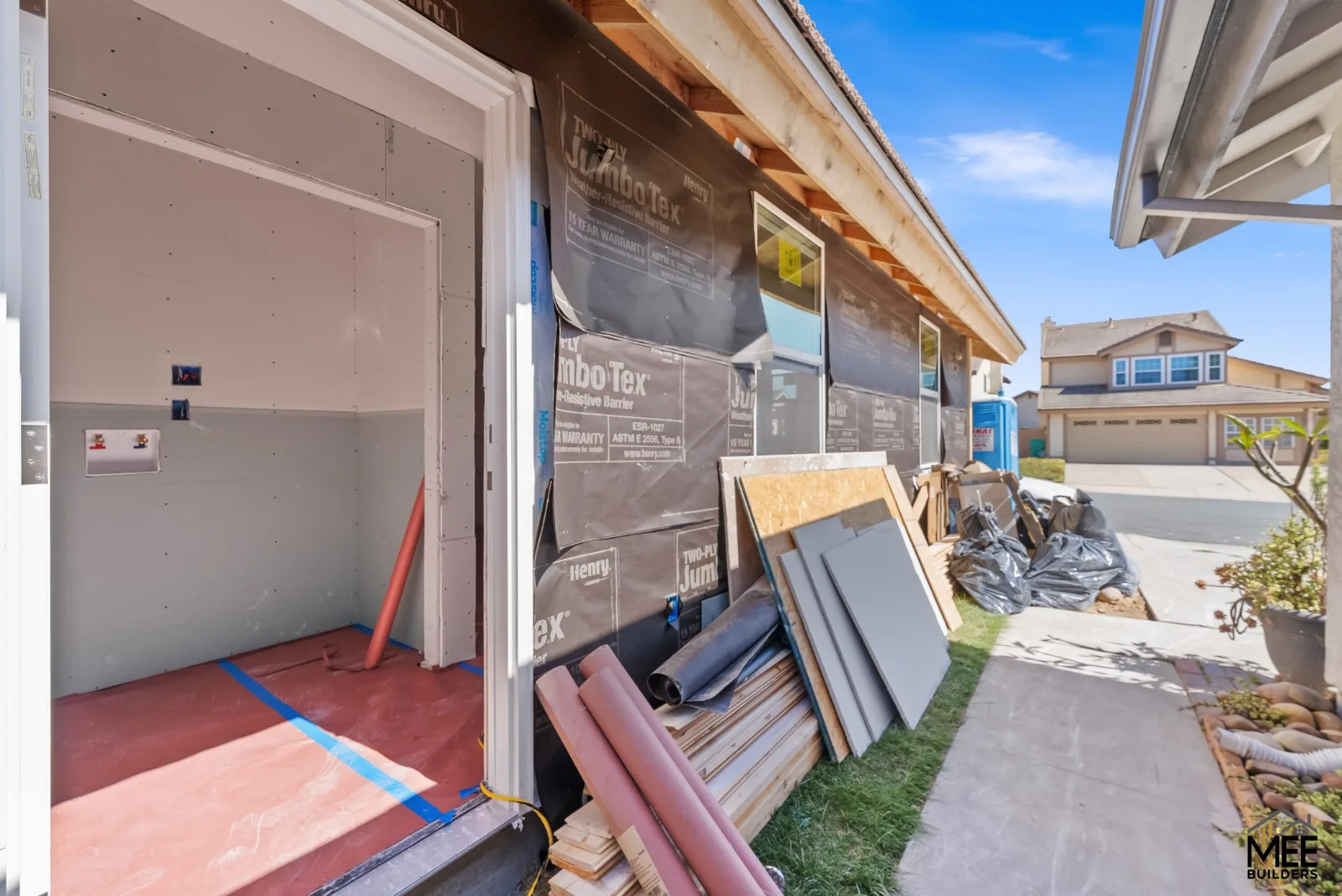 Unfinished exterior with wall wrap, exposed roof-edge framing, an open doorway, and construction materials beside a path.