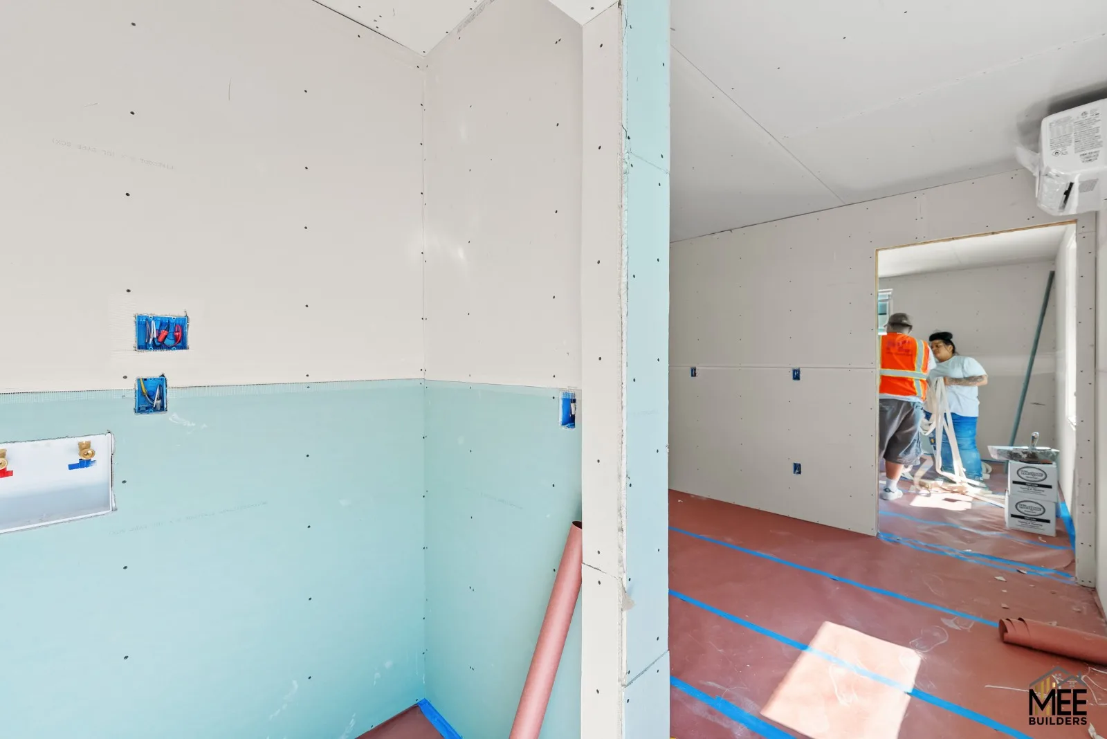Unfinished drywall interior with exposed electrical boxes, protected floors, and two workers through a doorway.