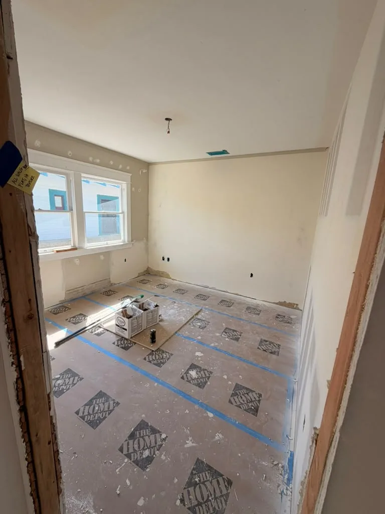 Unfinished room with patched drywall, two windows, ceiling wiring, and floor protection secured with blue tape.
