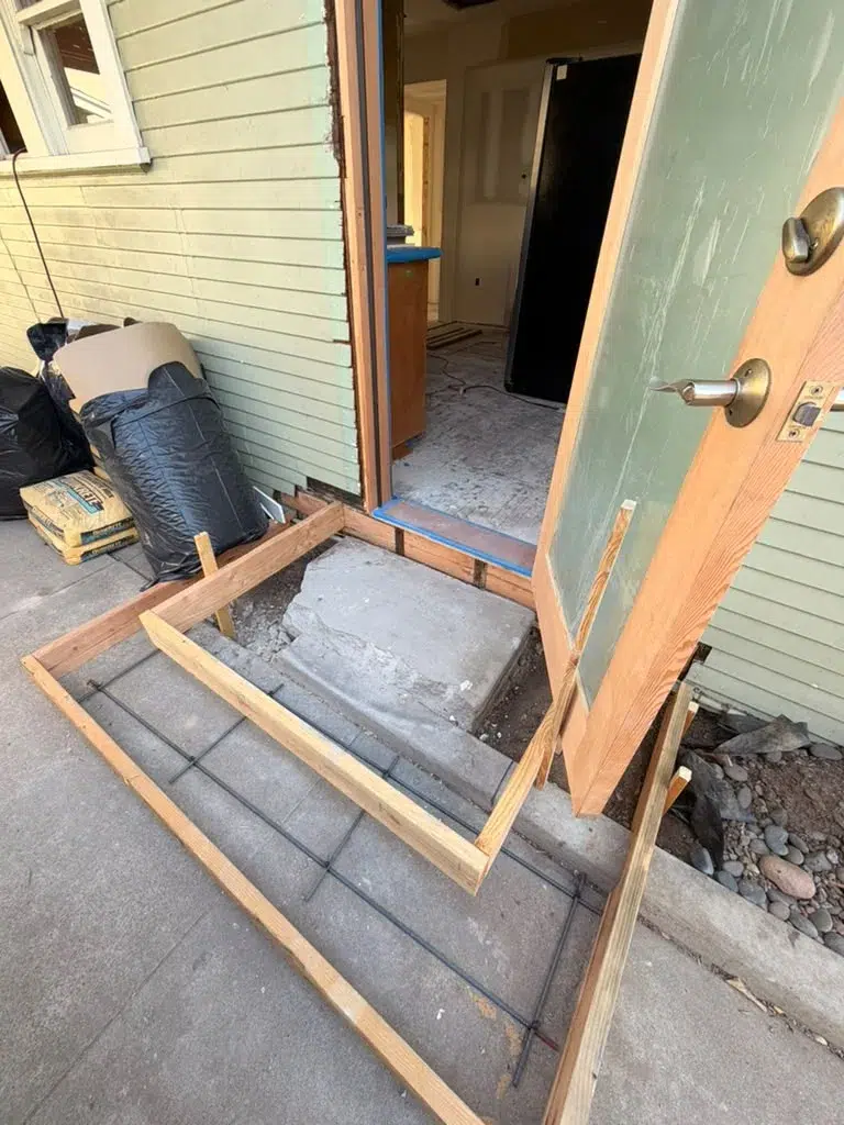 Open wood door above an entry step surrounded by lumber forms and exposed steel reinforcing bars.