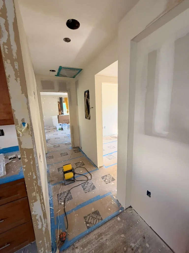Hallway with taped drywall seams, ceiling openings, protected floors, and a yellow work light.