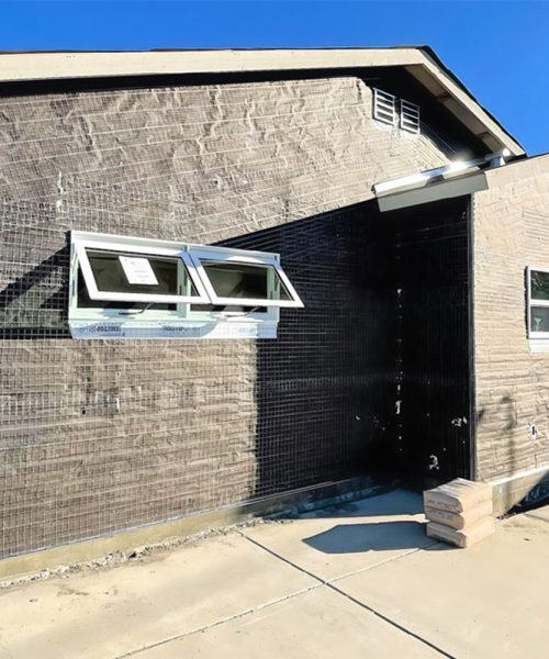 A modern single-story residential building under construction featuring a striking black vertical accent wall contrasted with light tan brick. Two awning-style windows are prominently installed on the dark section, and a side entrance with a door is visible on the right portion of the structure. The building sits on a concrete foundation with a clear walkway, photographed against a bright blue sky.