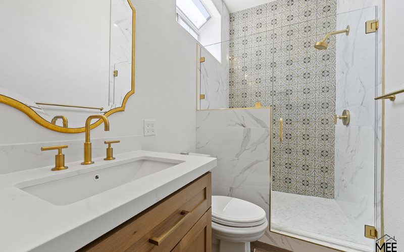 A modern bathroom featuring a wooden vanity with white countertop and gold fixtures, paired with a decorative gold-framed mirror. The shower area showcases patterned tile accent wall combined with white marble-look tiles and a frameless glass door with gold hardware. Natural light enters through a skylight above the shower.