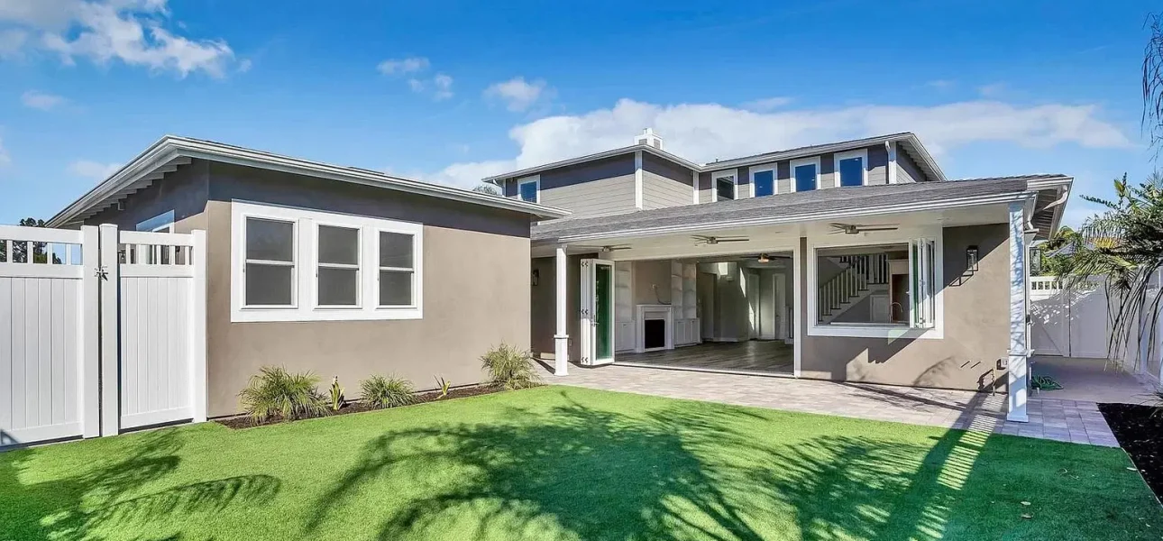A modern two-story residential home with beige and gray exterior siding features an open-concept design with large glass doors connecting indoor and outdoor spaces. The backyard showcases vibrant artificial green turf lawn with a covered patio area and white-trimmed garage or storage structure on the left side. The contemporary architecture includes clean lines, multiple windows, and a second-story section visible above the main level under a clear blue sky with scattered clouds.