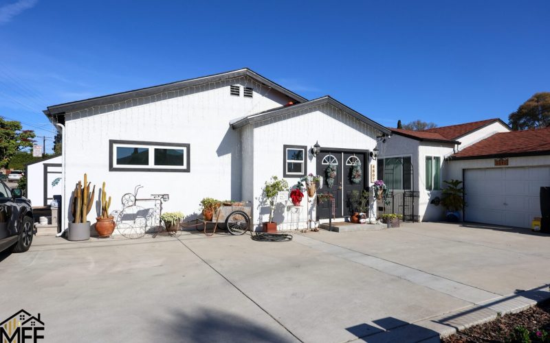 A white stucco single-family home with dark-trimmed windows and decorative gabled rooflines sits on a concrete driveway under a bright blue sky. The front yard features various potted plants, cacti, decorative items including a bicycle and wreath-adorned entrance. Adjacent homes are visible on either side, with a garage structure to the right, showcasing a typical residential neighborhood setting.