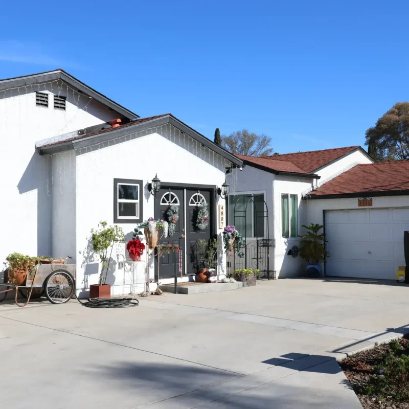 A single-story white residential property with multiple connected structures featuring gray-trimmed roofing and arched doorway details. The home has a wide concrete driveway leading to an attached garage, with decorative elements including potted plants, wreaths, and a vintage-style bicycle near the entrance. The property is set against a clear blue sky with mature trees visible in the background.
