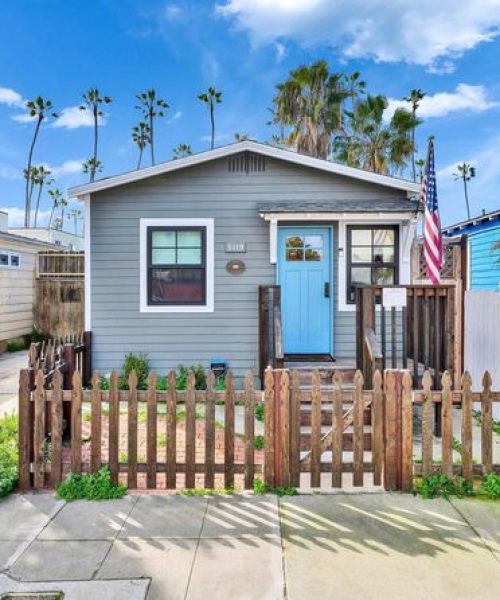 A small single-story beach cottage with light blue-gray siding and white trim, featuring a front porch with an American flag. The home is enclosed by a rustic wooden picket fence and surrounded by tall palm trees under a bright blue sky with white clouds. The coastal property has a casual, charming beach community aesthetic typical of Southern California seaside neighborhoods.