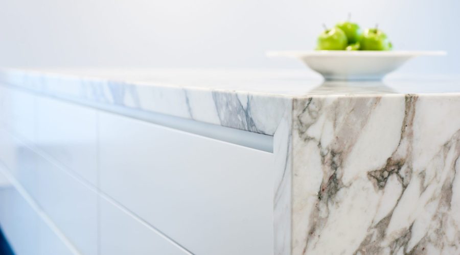 A modern kitchen detail showing the edge of a marble countertop with natural brown and gray veining, white cabinet drawers below, and a shallow white bowl filled with green apples positioned on the marble surface. The clean, minimalist design emphasizes the contrast between the sleek white cabinetry and the organic patterns in the natural stone countertop.