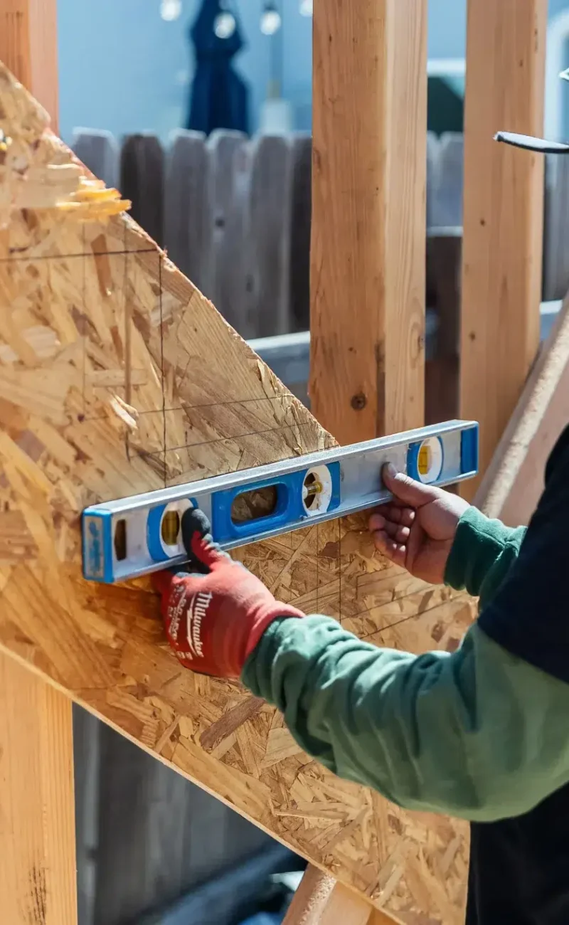 A construction worker wearing a green hood and work gloves uses a blue spirit level to check the alignment of oriented strand board (OSB) sheathing on a wooden frame structure. The worker is positioned between wooden studs, carefully ensuring the angled roof or wall panel is properly leveled during the building process. The construction site appears to be an active outdoor project with framing visible in the background.