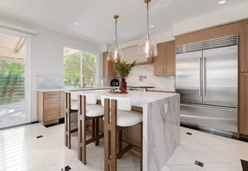 A modern kitchen featuring light wood cabinetry and a large marble waterfall island with three white upholstered bar stools. Two glass pendant lights hang above the island, which displays decorative items including flowers and a coffee maker. The space includes stainless steel appliances, white tile flooring with black accent tiles, and large windows overlooking greenery.