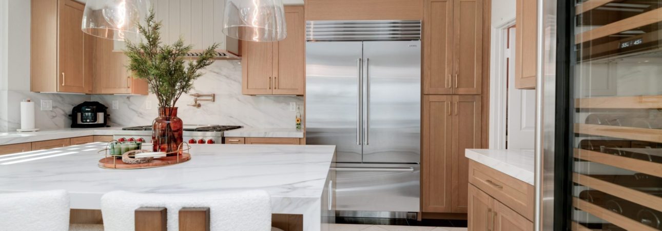 A modern kitchen featuring light wood cabinetry paired with white marble countertops and backsplash. The space includes a central island with white upholstered bar stools, glass pendant lights, a stainless steel refrigerator, and a wine storage unit visible on the right. The design combines warm wood tones with white surfaces and metallic accents in a clean, contemporary style.