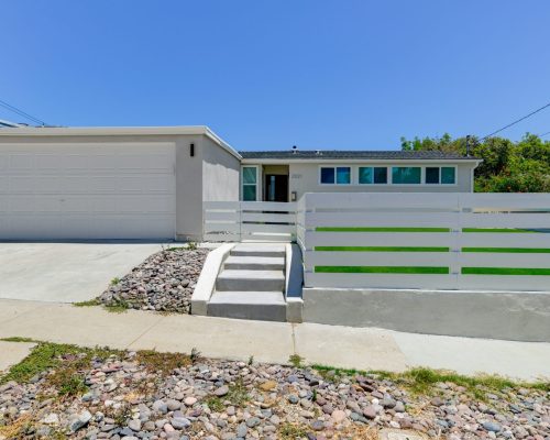 A single-story ranch-style home with white exterior walls and a two-car garage, featuring a horizontal white fence with green accent stripes along the front. The property has concrete steps leading up from the sidewalk, with drought-resistant landscaping consisting of river rock beds on either side of the walkway. Several turquoise-colored windows are visible across the front facade under a clear blue sky.