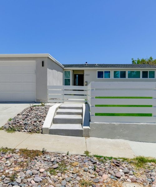 A single-story ranch-style home with white exterior walls and a two-car garage, featuring a horizontal white fence with green accent stripes along the front. The property has concrete steps leading up from the sidewalk, with drought-resistant landscaping consisting of river rock beds on either side of the walkway. Several turquoise-colored windows are visible across the front facade under a clear blue sky.