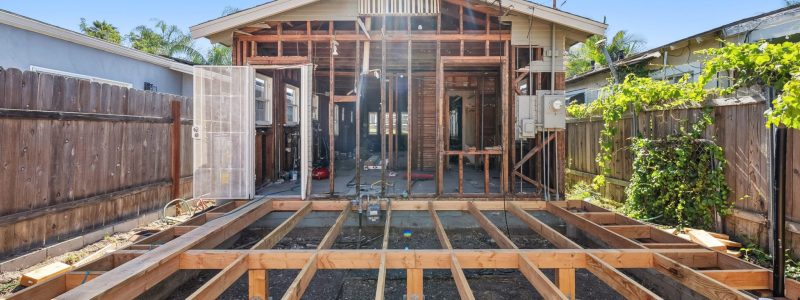 A residential construction site showing wooden floor joists and foundation framing in the foreground, leading to a house under renovation with exposed wall framing and a pitched roof. The structure is surrounded by wooden privacy fences and neighboring homes, with palm trees visible in the background under a clear blue sky. The MEE Builders logo appears in the bottom right corner.