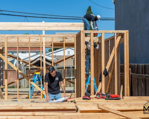 Construction workers frame a wooden structure on a bright sunny day. One worker stands on the floor working with materials while two others work on the top beam and wall frame. The setting shows a residential construction site with power lines, fencing, and neighboring buildings visible in the background, with the MEE Builders logo in the bottom right corner.