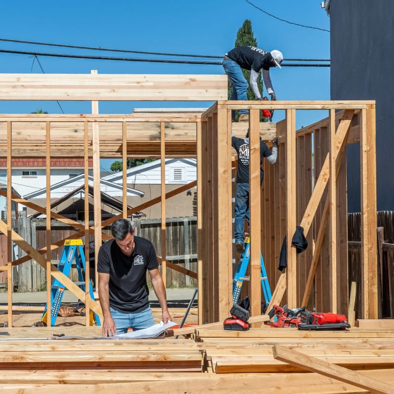 Construction workers frame a wooden structure on a bright sunny day. One worker stands on the floor working with materials while two others work on the top beam and wall frame. The setting shows a residential construction site with power lines, fencing, and neighboring buildings visible in the background, with the MEE Builders logo in the bottom right corner.