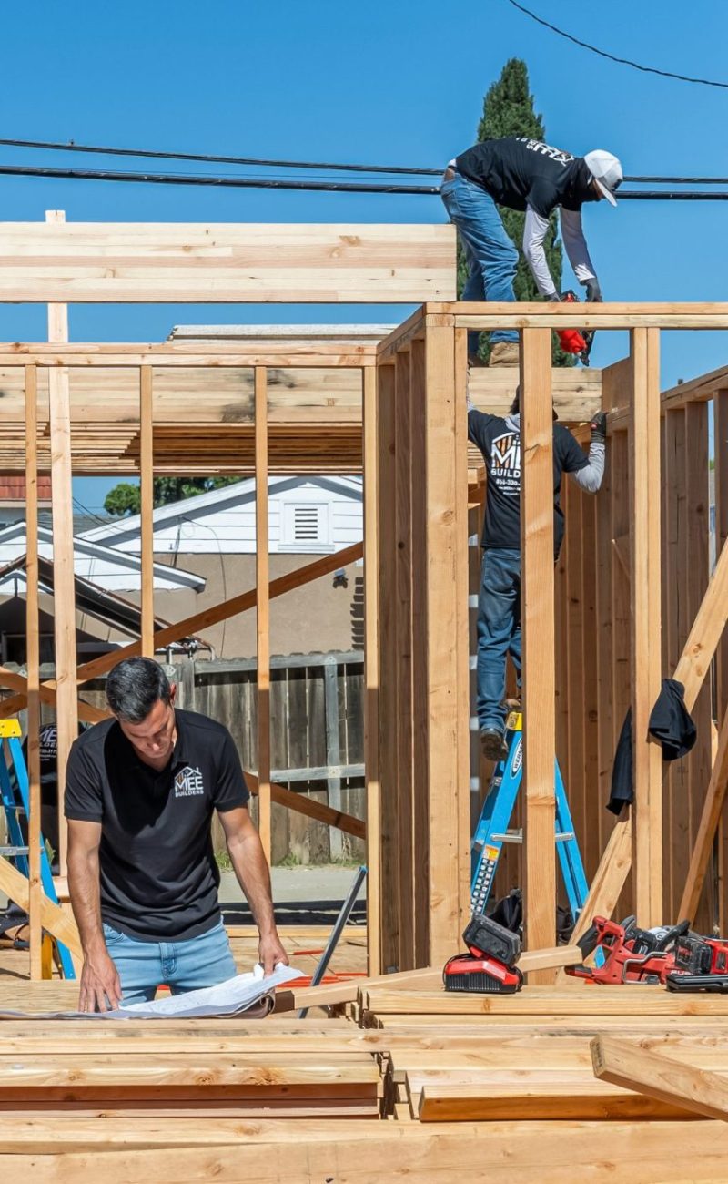 Construction workers frame a wooden structure on a bright sunny day. One worker stands on the floor working with materials while two others work on the top beam and wall frame. The setting shows a residential construction site with power lines, fencing, and neighboring buildings visible in the background, with the MEE Builders logo in the bottom right corner.