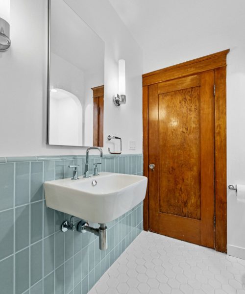 A bright bathroom featuring white hexagonal floor tiles and pale blue vertical subway tiles on the lower wall. A white wall-mounted sink with chrome fixtures sits beneath a large frameless mirror, while a wooden door with visible grain stands against the white upper walls. The space has vaulted ceilings and combines classic design elements with a clean, minimalist aesthetic.