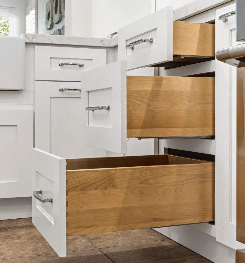 A modern white kitchen featuring multiple pull-out drawers in various sizes, showcasing natural wood interiors with dovetail joinery. The cabinets display soft-close drawer systems with sleek brushed nickel handles, demonstrating high-quality storage solutions and organizational features typical of custom cabinetry by MEE Builders.