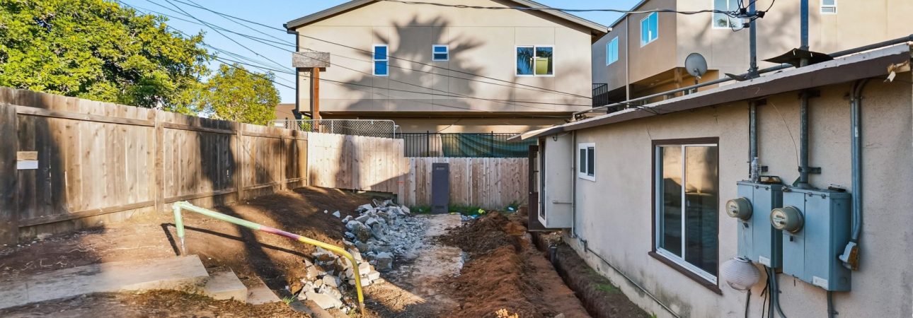 A residential construction site showing a backyard area with exposed dirt and excavation work in progress. The yard is bordered by wooden fences and surrounded by neighboring multi-story homes. Fresh soil grading and utility work are visible, with electric meters mounted on the right-side structure under a clear blue sky.