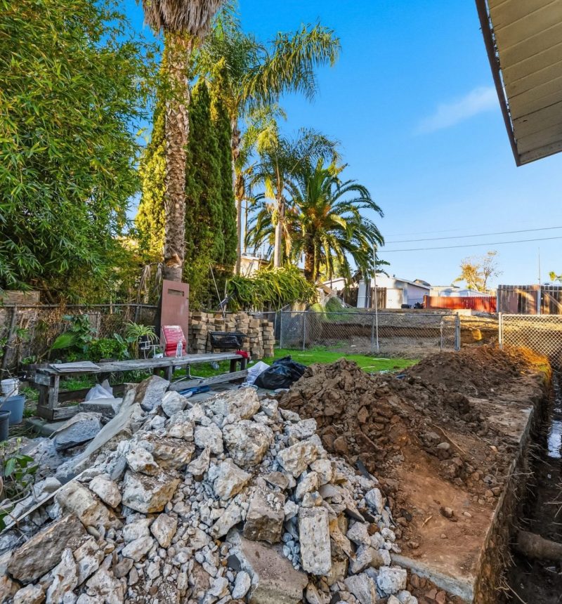 A residential backyard construction site showing excavated soil and broken concrete debris piled near the house foundation. Palm trees and lush green vegetation are visible in the background, with a chain-link fence separating the property from neighboring homes under a clear blue sky. The MEE Builders logo appears in the bottom right corner of the image.