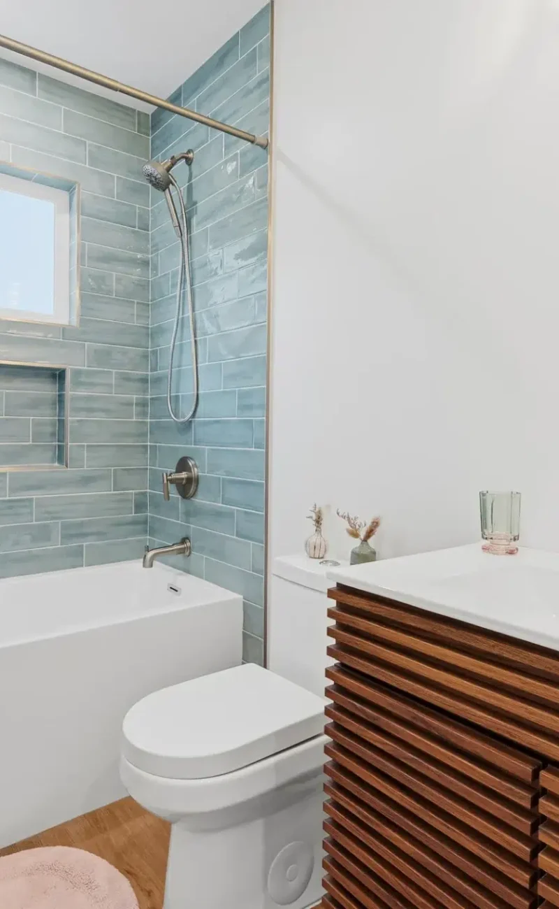 A modern bathroom featuring a white bathtub with aqua blue subway tile surround extending to the ceiling. The shower area includes a recessed niche and window for natural light. A contemporary vanity with horizontal wood slat detailing and white countertop sits alongside a white toilet, with brushed nickel fixtures throughout.