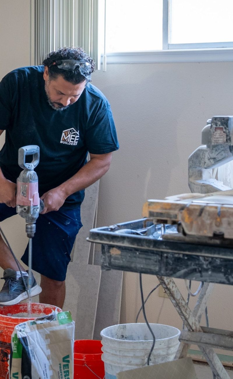 A construction worker in a dark blue t-shirt and shorts uses a mixing drill to blend materials in a red bucket. Behind him is a yellow tile saw on a work table covered with protective plastic sheeting. The room appears to be under renovation with bare walls and windows visible in the background.