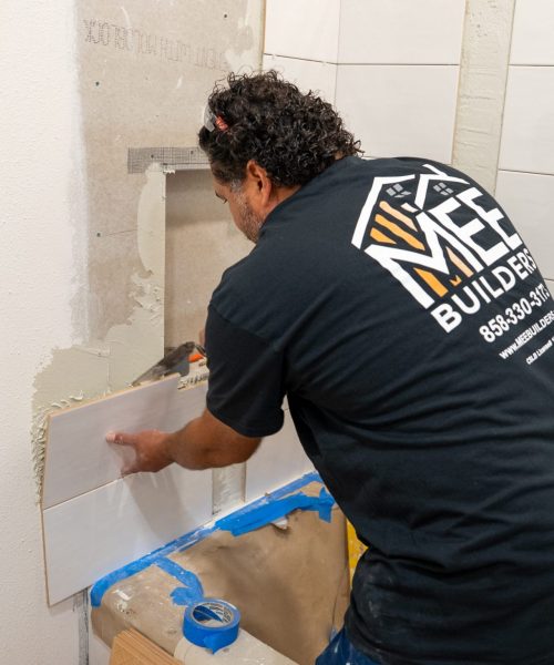 A worker from MH Builders wearing a black company shirt installs large white rectangular tiles on a bathroom wall. The person is positioned near a recessed shower niche, with blue painter's tape protecting the floor and surrounding areas. The wall shows partially completed tile work with visible adhesive and mesh backing material.