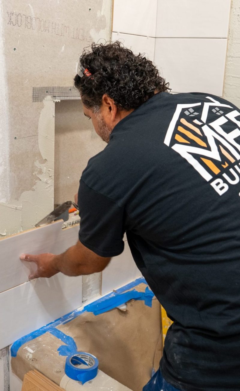 A worker from MH Builders wearing a black company shirt installs large white rectangular tiles on a bathroom wall. The person is positioned near a recessed shower niche, with blue painter's tape protecting the floor and surrounding areas. The wall shows partially completed tile work with visible adhesive and mesh backing material.