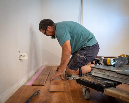 A man in a teal shirt and dark shorts kneels on a wooden floor while installing laminate flooring planks. He's working near a white wall with an electrical outlet, using tools including what appears to be a power drill visible on a work surface beside him. The flooring installation is in progress, with several planks already laid and green painter's tape visible along the wall edge.