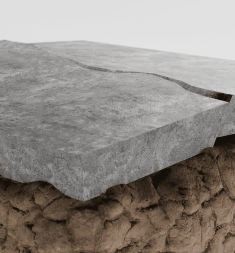 A three-dimensional cross-section showing two gray concrete slabs positioned above a textured brown substrate layer. The concrete pieces display a visible crack or gap between them, illustrating how concrete surfaces can separate or shift. The brown base layer appears rough and uneven, representing the ground or underlying material beneath a concrete foundation or pavement.