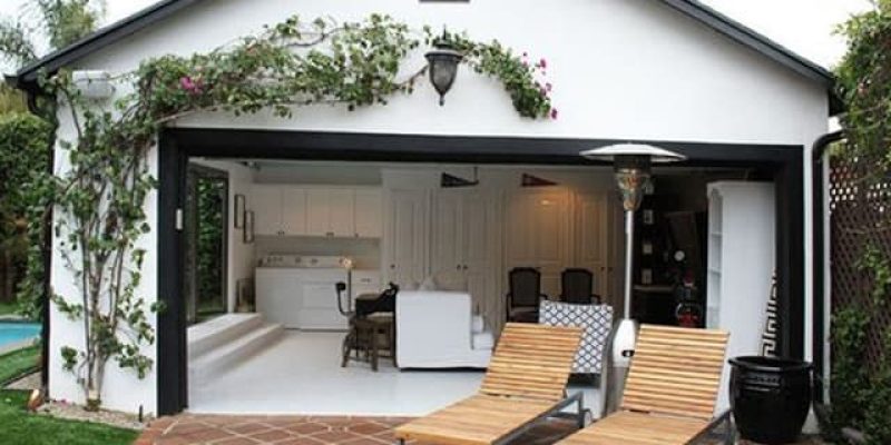 A modern white garage conversion with large black-framed opening showcasing a bright interior living space. Two wooden lounge chairs sit on a curved terracotta tile patio in front of the garage, while climbing vines and flowers adorn the exterior walls. The interior features white walls, modern furniture including chairs and seating, creating a seamless indoor-outdoor living area.
