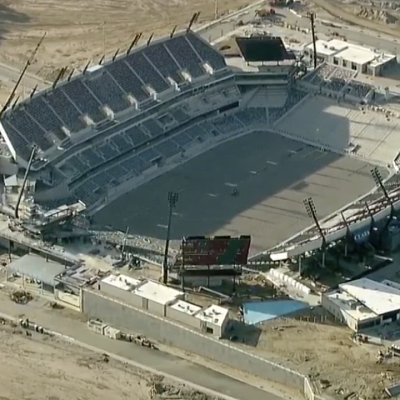Aerial view of a football stadium under construction, showing a horseshoe-shaped seating bowl with one completed side featuring multiple tiers and a partially constructed opposite side with exposed steel framework. The facility is surrounded by barren land and construction equipment, with parking areas and roadways visible in the background. The dark playing field surface is already installed within the stadium structure.