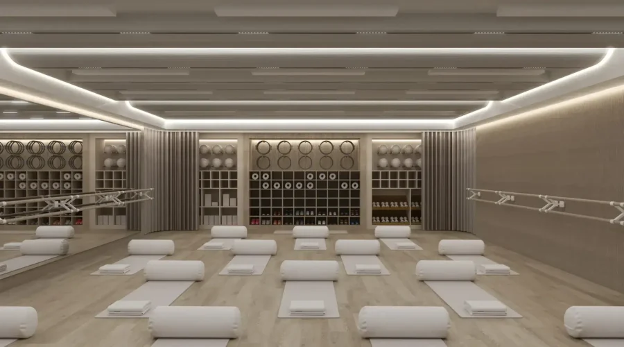 A modern meditation or yoga studio with rows of white cushioned bolsters and mats arranged on a light wood floor. The minimalist space features recessed ceiling lighting with linear LED strips, neutral beige walls with decorative geometric panels, and built-in shelving units displaying meditation props and accessories along the back wall.